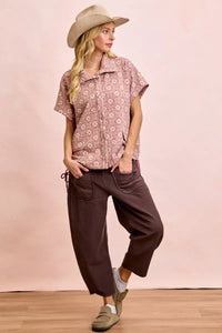 Woman wearing a patterned shirt and brown pants against a pink background