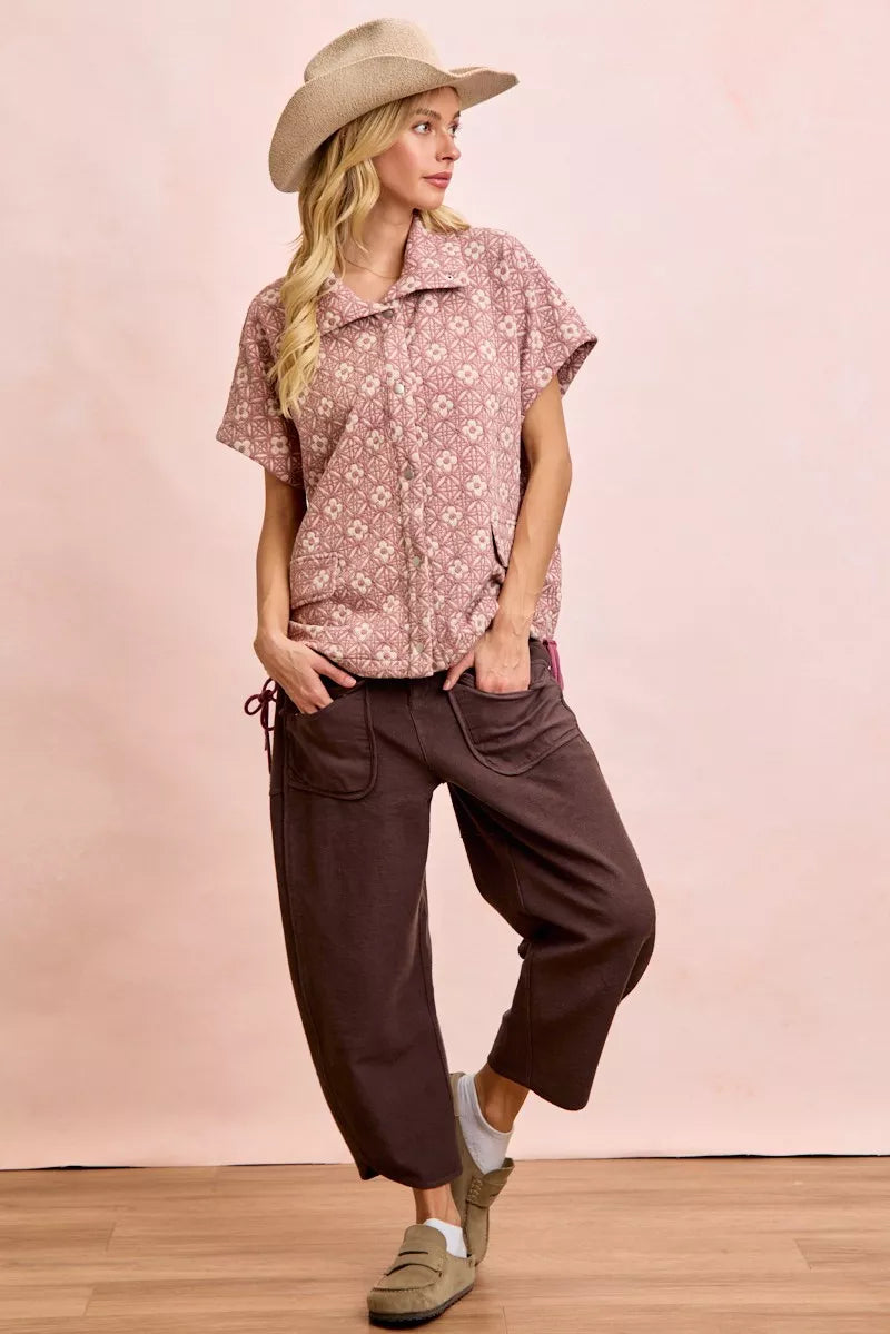Woman wearing a patterned shirt and brown pants against a pink background