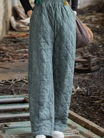 A person wearing quilted wide-leg pants in a muted sage green color, standing outdoors on a weathered wooden platform with a rustic, cluttered background.