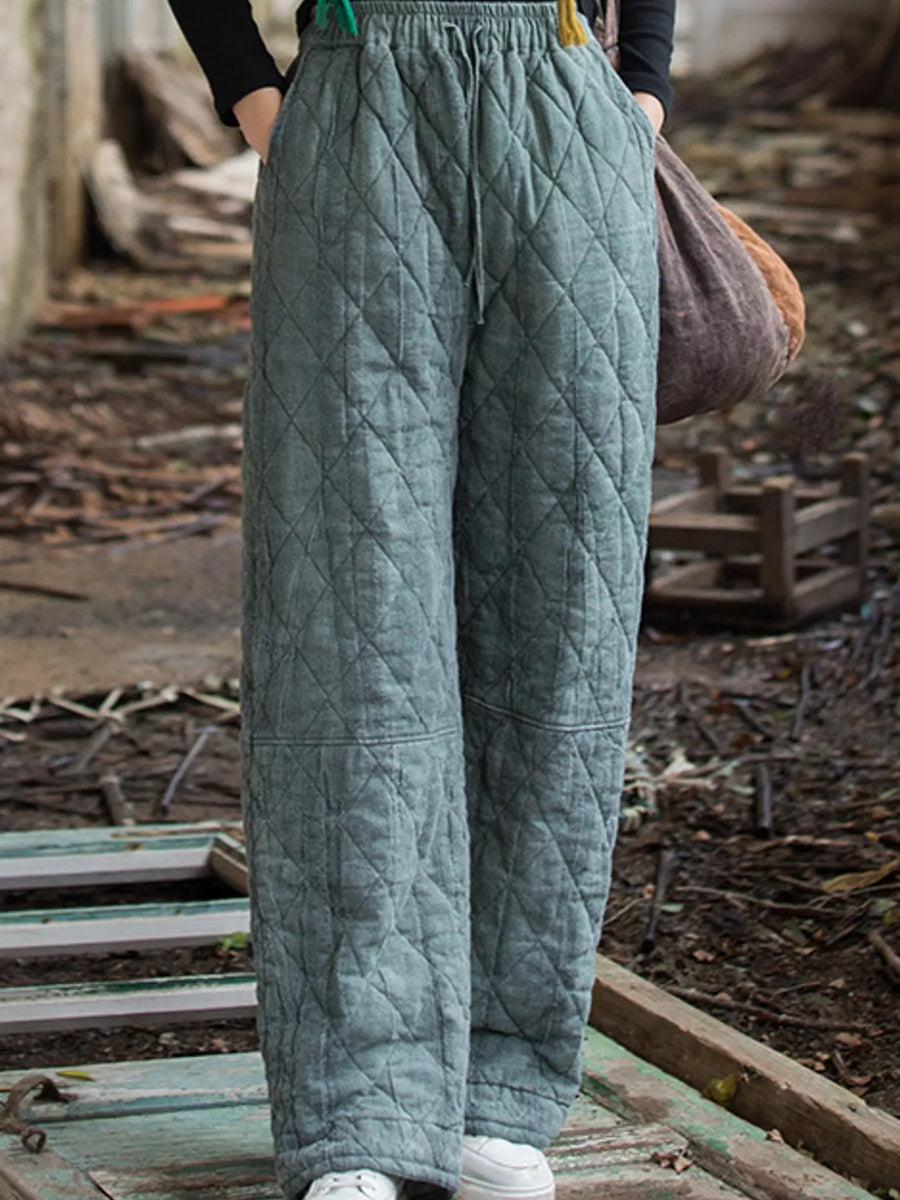 A person wearing quilted wide-leg pants in a muted sage green color, standing outdoors on a weathered wooden platform with a rustic, cluttered background.