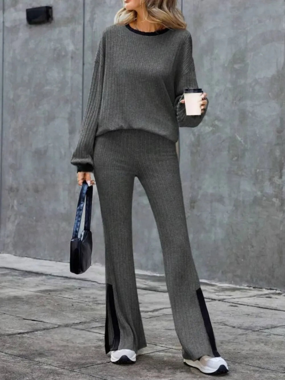 Person wearing a gray outfit with a black handbag and white shoes, standing against a gray wall.