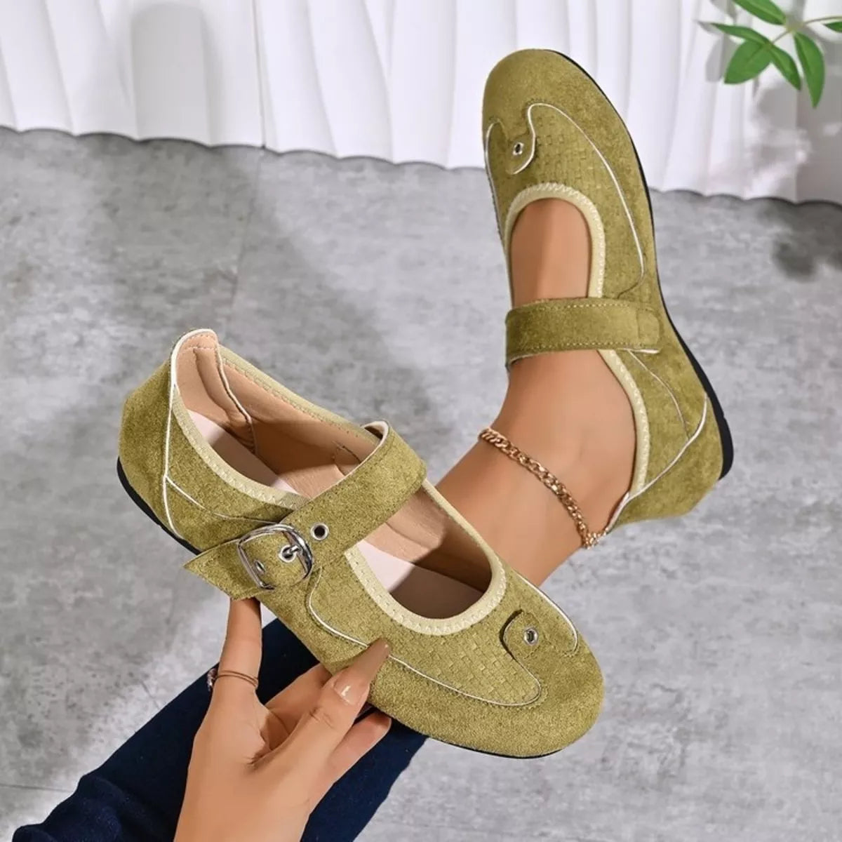 A pair of women's low-heeled Mary Jane shoes in olive green suede, shown being worn and held by a hand. The shoes feature a strap with a silver buckle, decorative stitched details, and small bead accents.