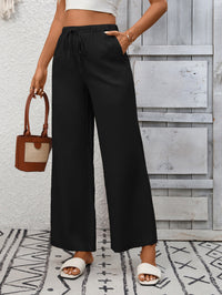 Person wearing black wide-leg pants and white sandals, holding a brown handbag against a wooden panel background.