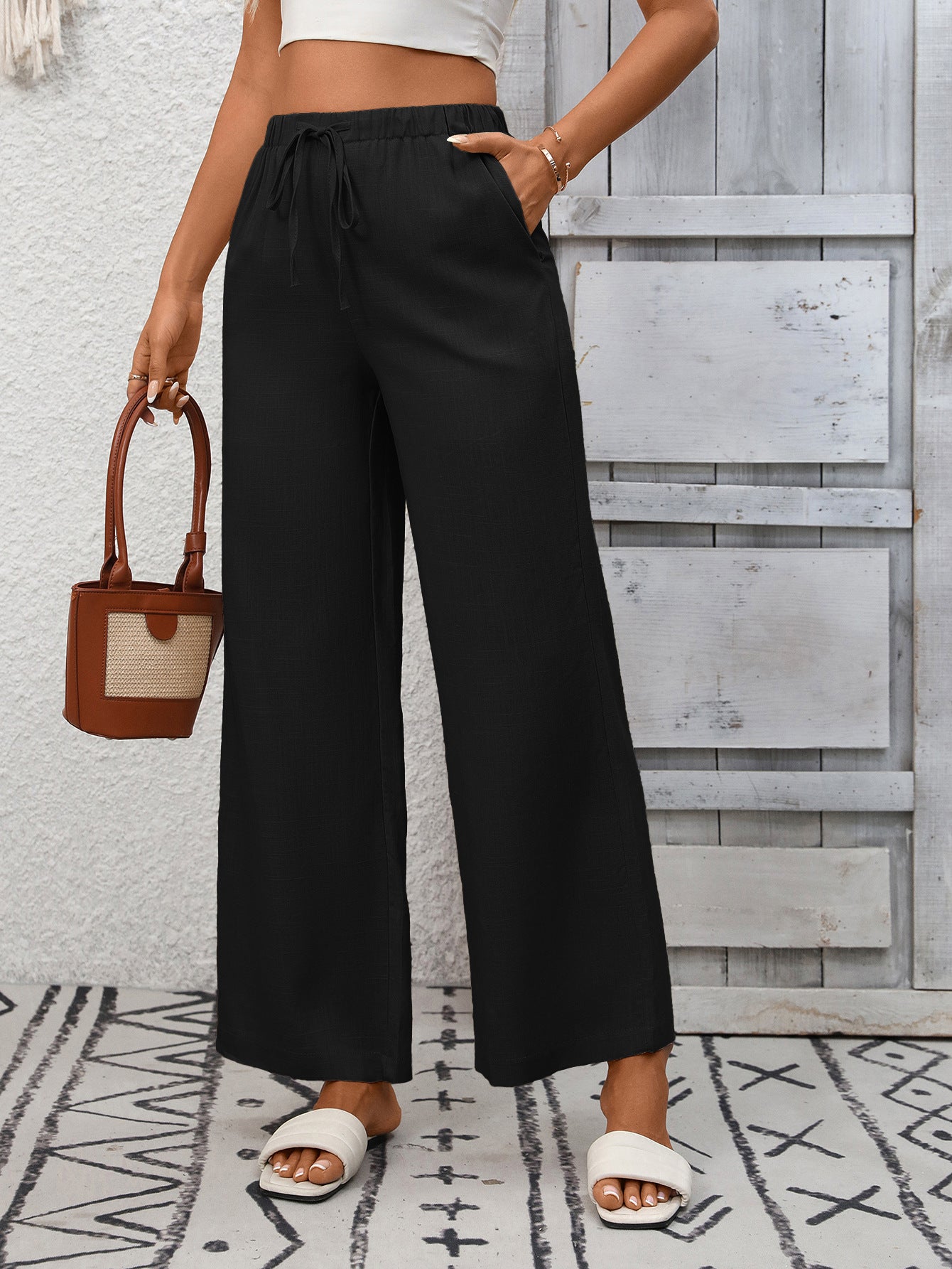 Person wearing black wide-leg pants and white sandals, holding a brown handbag against a wooden panel background.