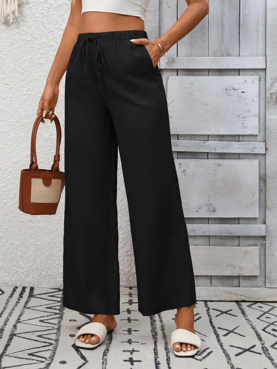 Person wearing black wide-leg pants and white sandals, holding a brown handbag against a wooden panel background.