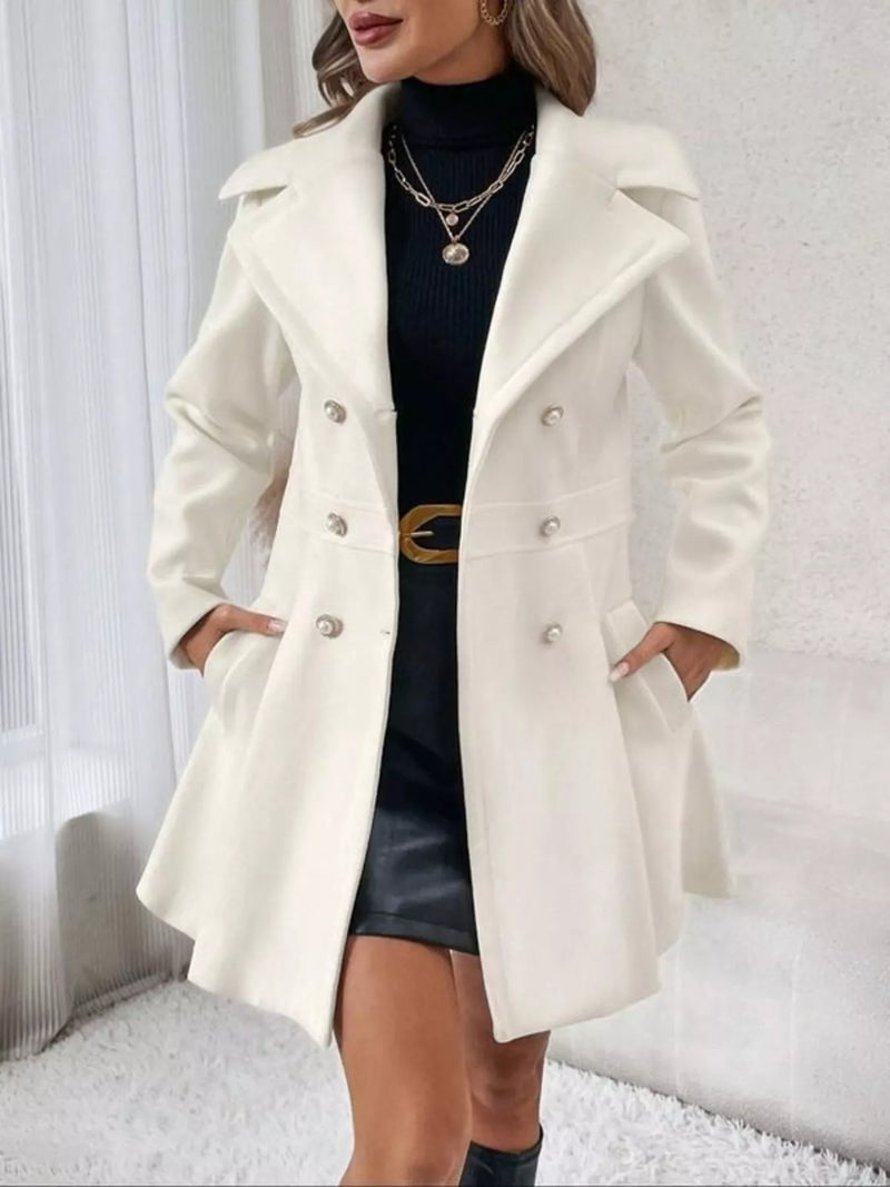A woman wearing a cream-colored double-breasted coat over a black turtleneck and black skirt, standing in a bright indoor setting with white walls and a white rug.