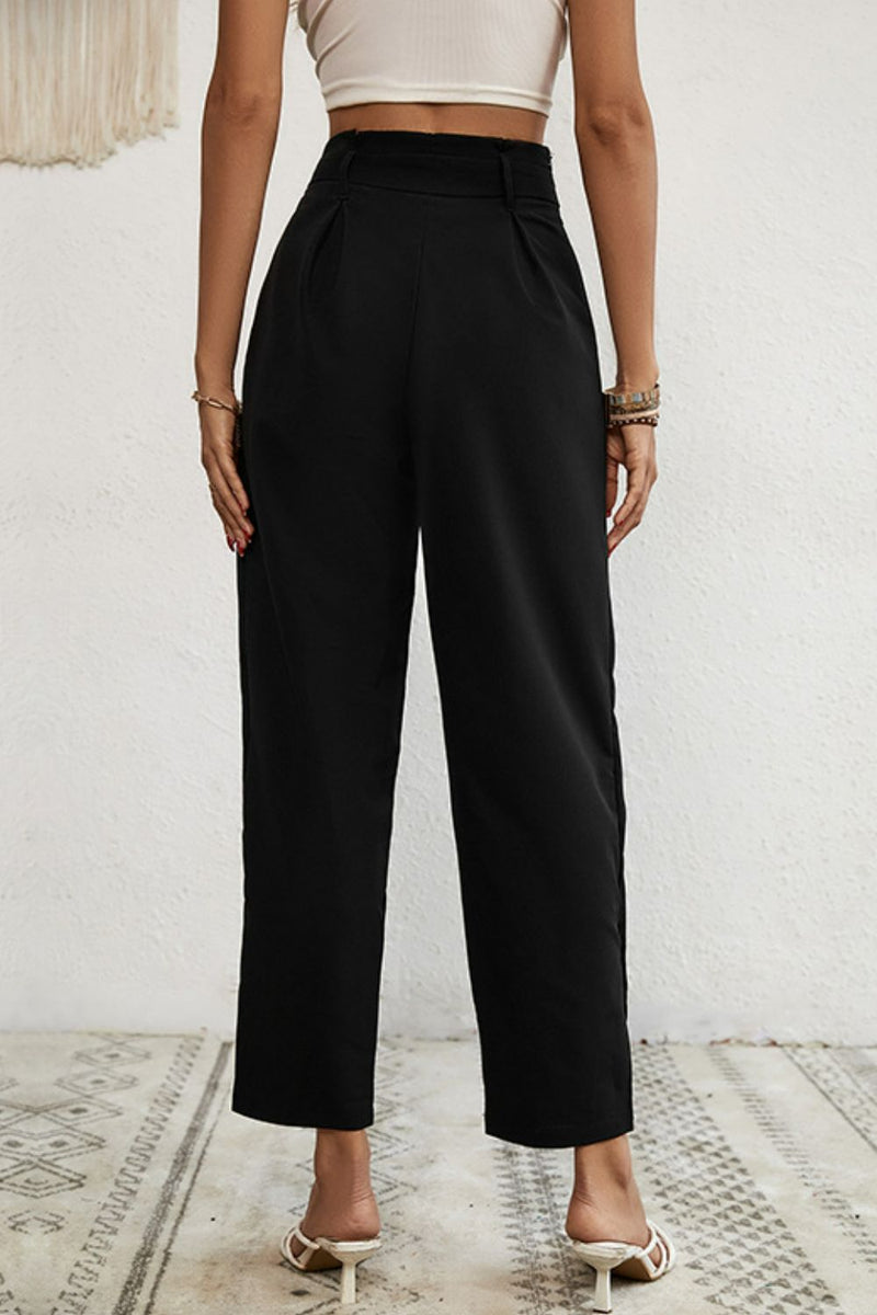 Person wearing black high-waisted pants and a beige top against a white wall.