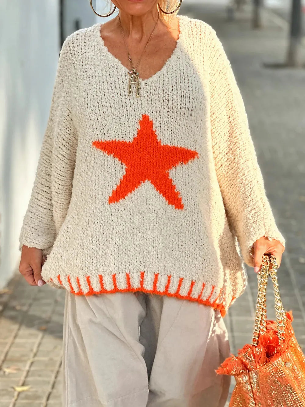 Person wearing a beige sweater with a red star design, holding an orange bag.