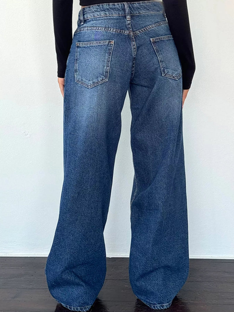 A person viewed from the back wearing loose-fit medium-wash blue denim jeans with a black long-sleeve top, standing against a plain white wall.
