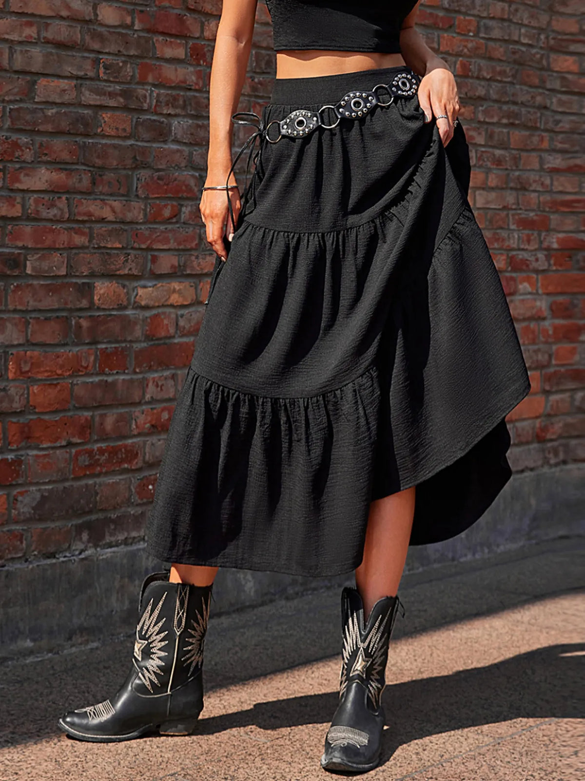 Person wearing a black skirt with decorative belt and black boots against a brick wall.