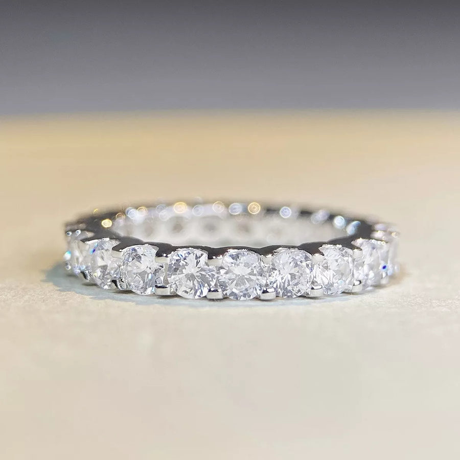 A silver diamond eternity ring with a row of round brilliant-cut diamonds set in a polished silver band, resting on a light beige surface with a soft gray background.