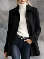 A woman wearing a black mid-length coat over a white ribbed turtleneck sweater, dark blue jeans, and a black belt with gold hardware, photographed against a soft gray blurred background.