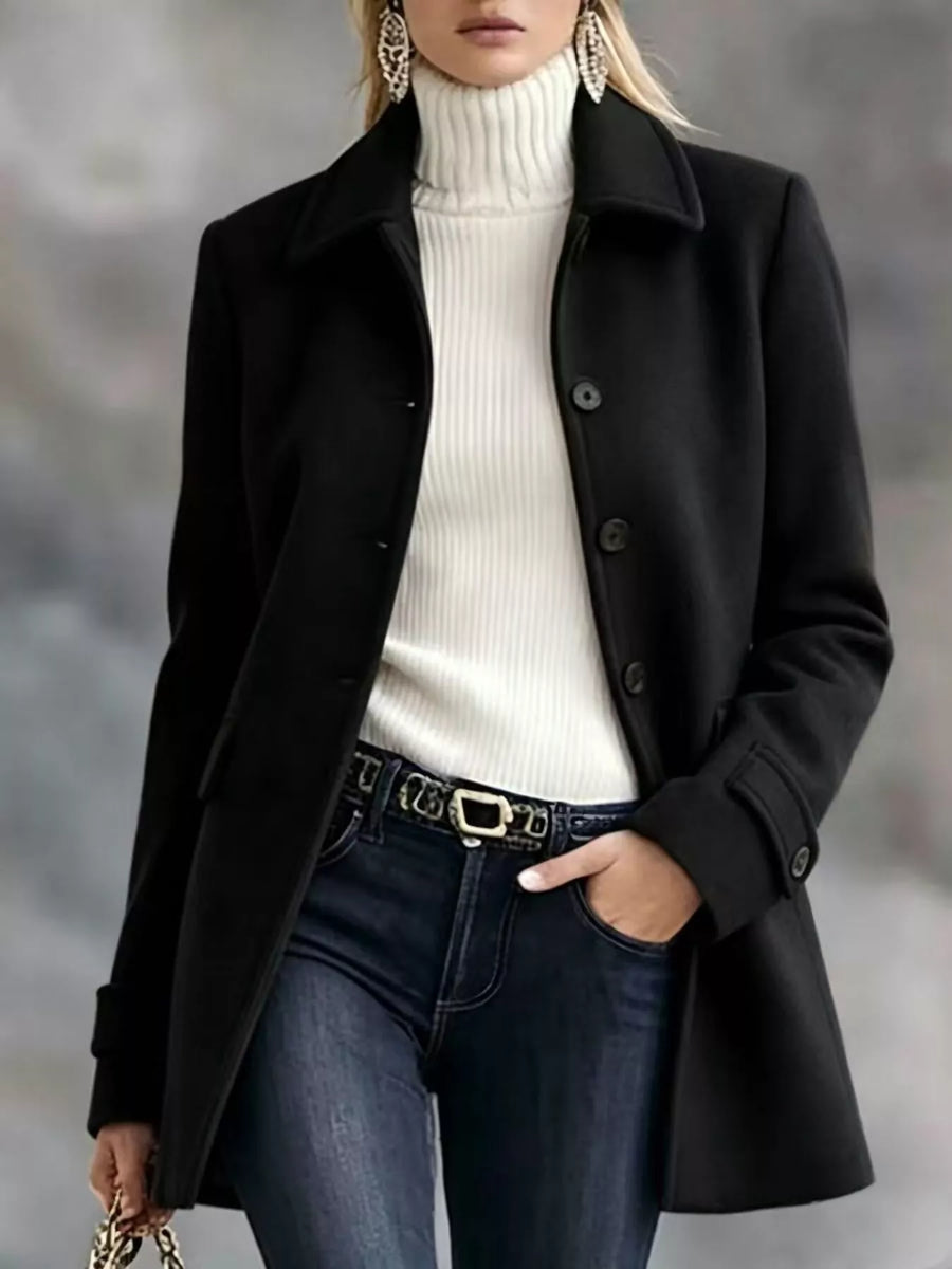 A woman wearing a black mid-length coat over a white ribbed turtleneck sweater, dark blue jeans, and a black belt with gold hardware, photographed against a soft gray blurred background.