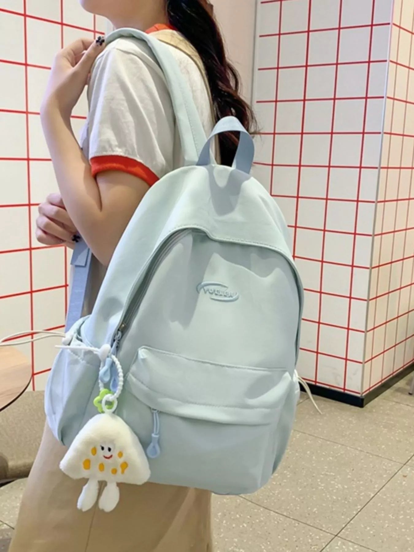 Person wearing a light blue backpack with a small white plush toy attached, against a red and white checkered wall.