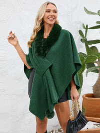 Woman wearing a green poncho with fur trim, holding a black handbag indoors.