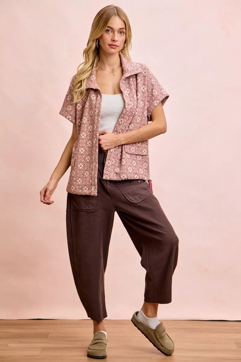 Woman wearing a patterned vest over a white top and brown pants on a pink background