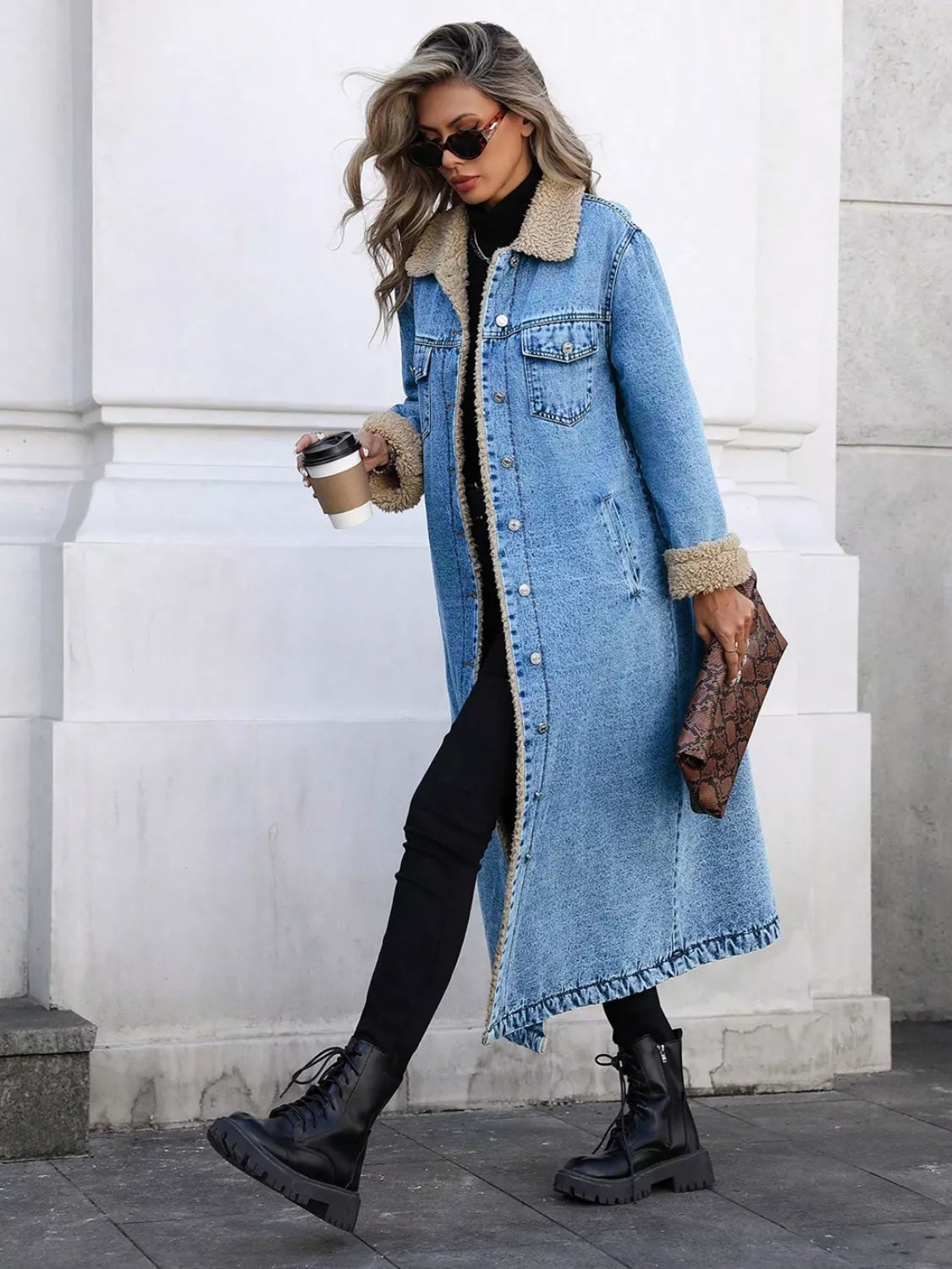 Woman wearing a long denim coat with fur trim, holding a coffee cup and clutch, standing against a white wall.
