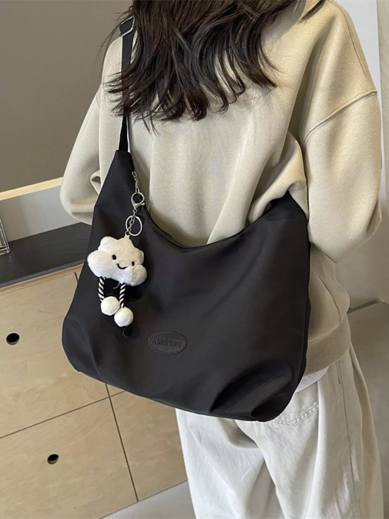 Person holding a black bag with a plush toy charm, wearing a beige sweater and light-colored pants.