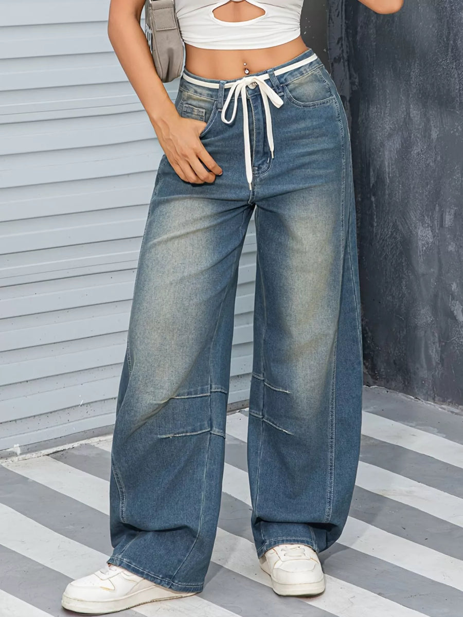 Person wearing wide-leg jeans with a white top and sneakers, standing against a striped floor and wall.