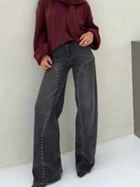 Person wearing maroon sweater and black jeans with studs on a white floor and wall.