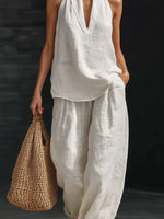 Person wearing a white sleeveless top and pants holding a woven bag against a dark background