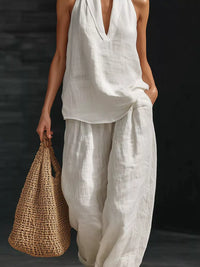 Person wearing a white sleeveless top and pants holding a woven bag against a dark background