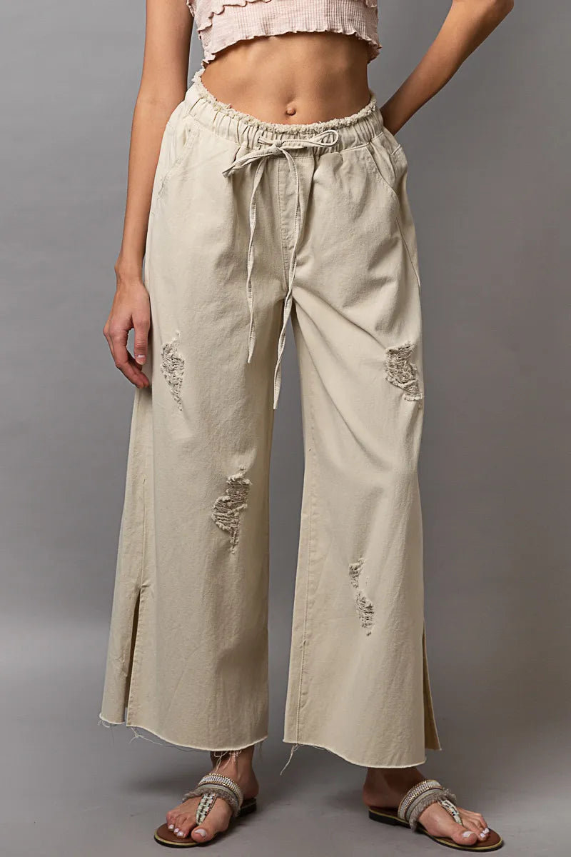 Beige wide-leg pants with distressed details worn by a person on a gray background