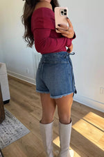 Person taking a mirror selfie wearing a red top, denim shorts, and white knee-high boots in a room with wooden flooring.