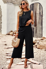 Woman in black sleeveless top and wide-leg pants standing on a stone path with rocks and a building in the background.