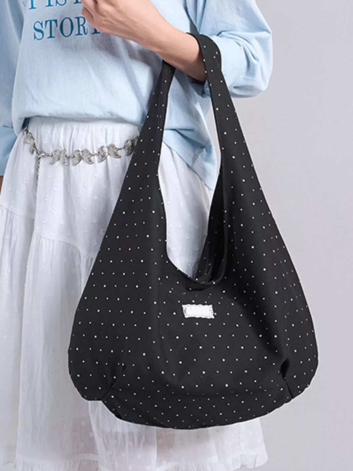Black polka dot bag held by a person wearing a light blue shirt and white skirt on a gray background