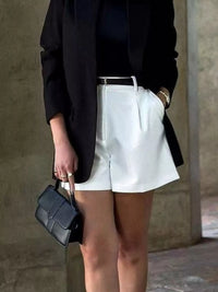 Person wearing a black blazer and white shorts holding a black handbag against a neutral background
