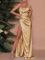 Woman wearing a gold satin gown with a high slit, standing against a beige background with green foliage.