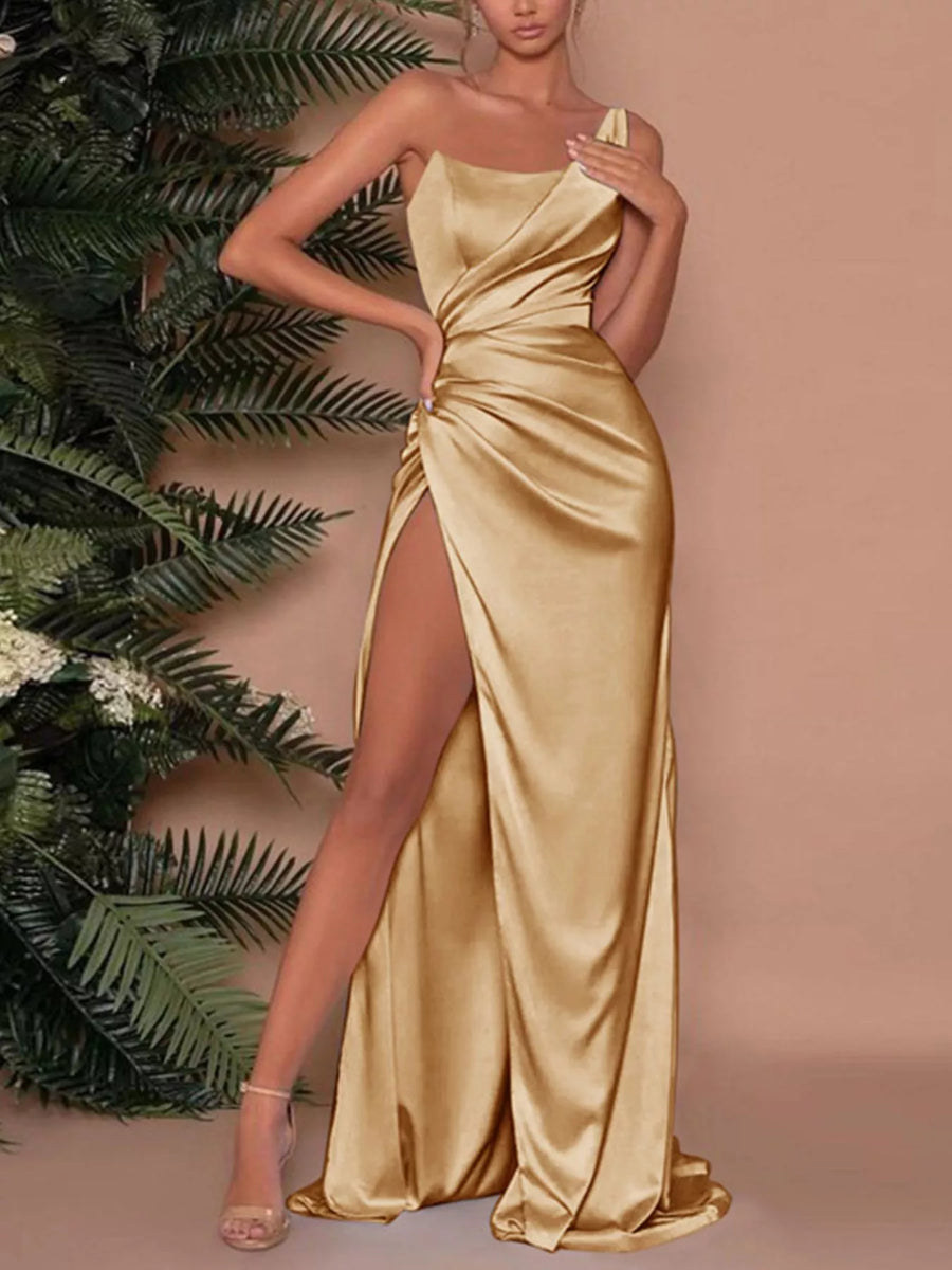 Woman wearing a gold satin gown with a high slit, standing against a beige background with green foliage.