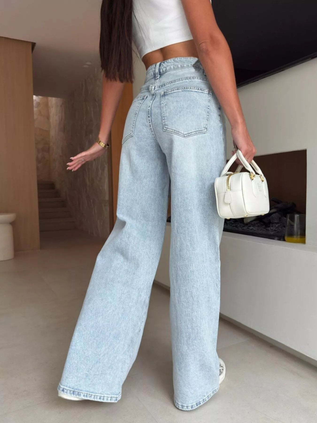 Person wearing light blue jeans and a white top, holding a small white handbag indoors.