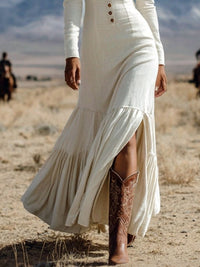 Person wearing a long white dress and brown boots in a desert setting with horses in the background.