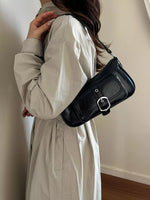 A woman wearing a light beige trench coat carries a black leather shoulder bag with a silver buckle, shown from a side-back angle against an indoor wall.