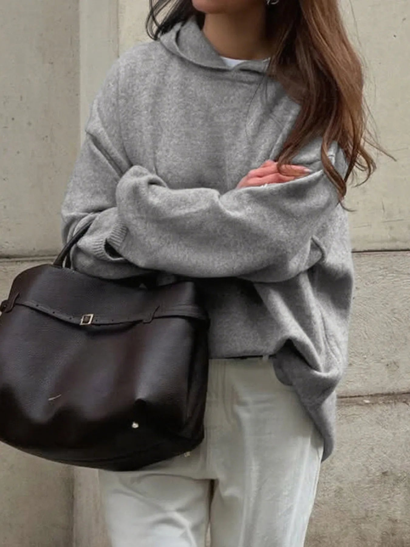 Person wearing a gray sweater and holding a brown leather bag against a neutral background