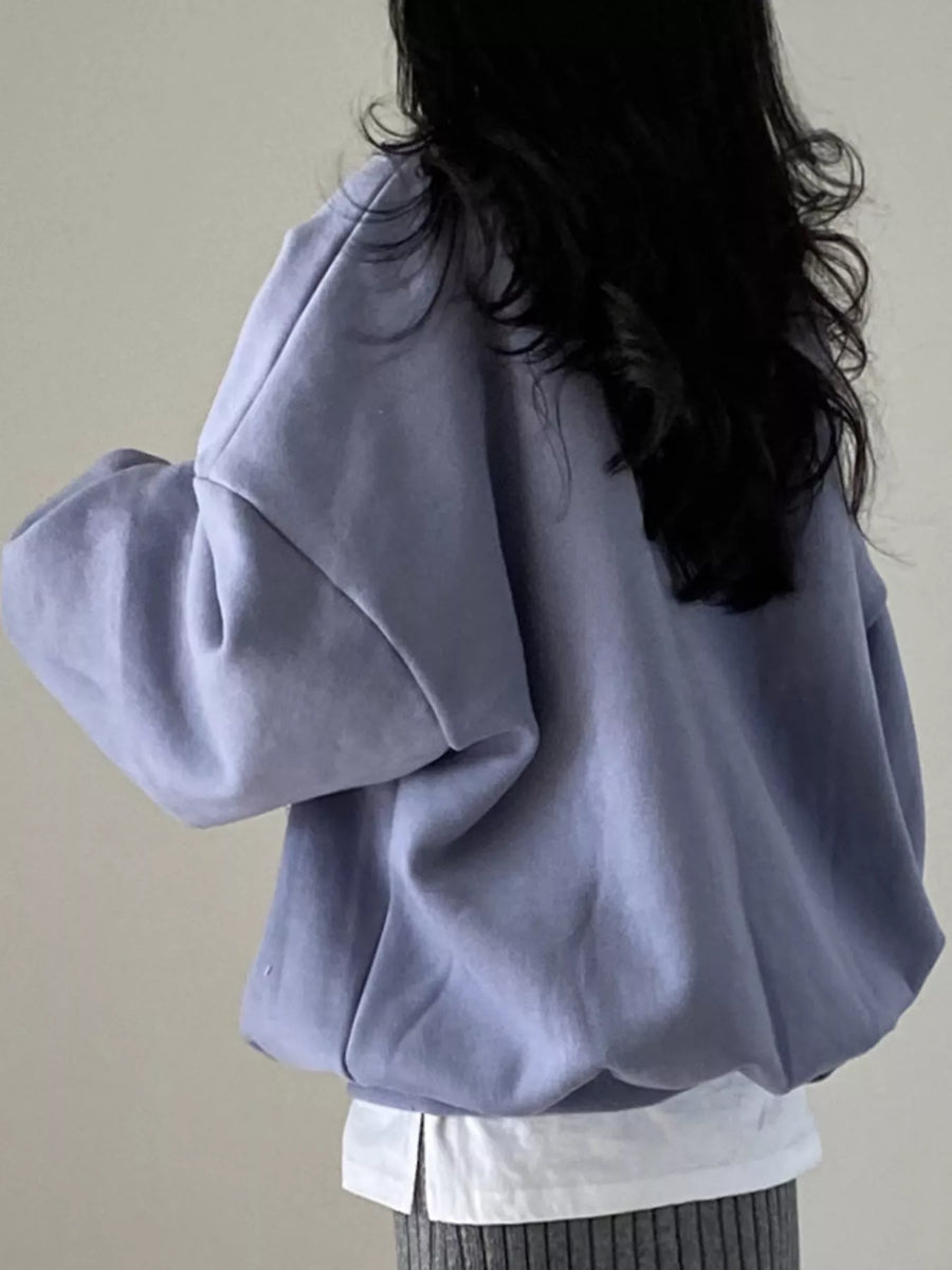 A person shown from the back wearing an oversized light lavender sweatshirt over a white shirt and gray ribbed pants, against a plain light beige wall.