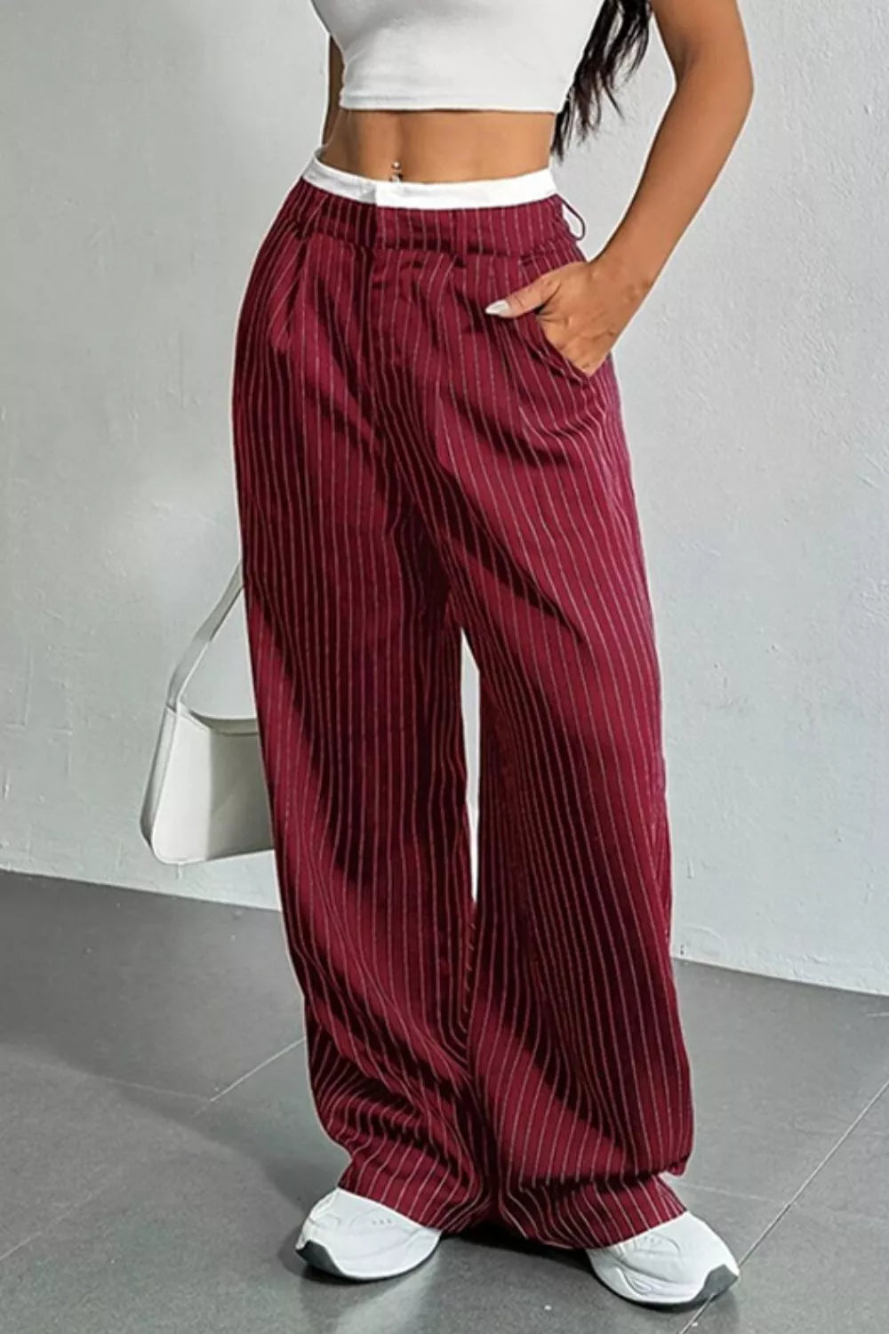 A person wearing loose, wide-leg burgundy pants with thin vertical white pinstripes, a white waistband, and side pockets. The model is also wearing a white crop top and white sneakers, standing against a light gray wall.