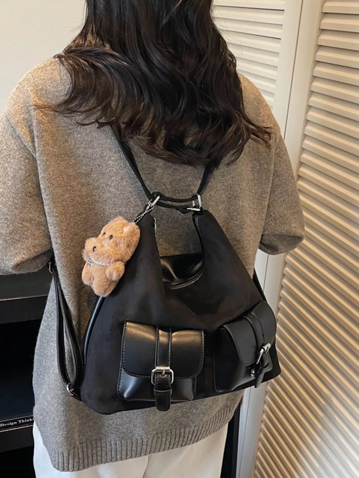 Person wearing a black backpack with a teddy bear charm, standing against a neutral background.