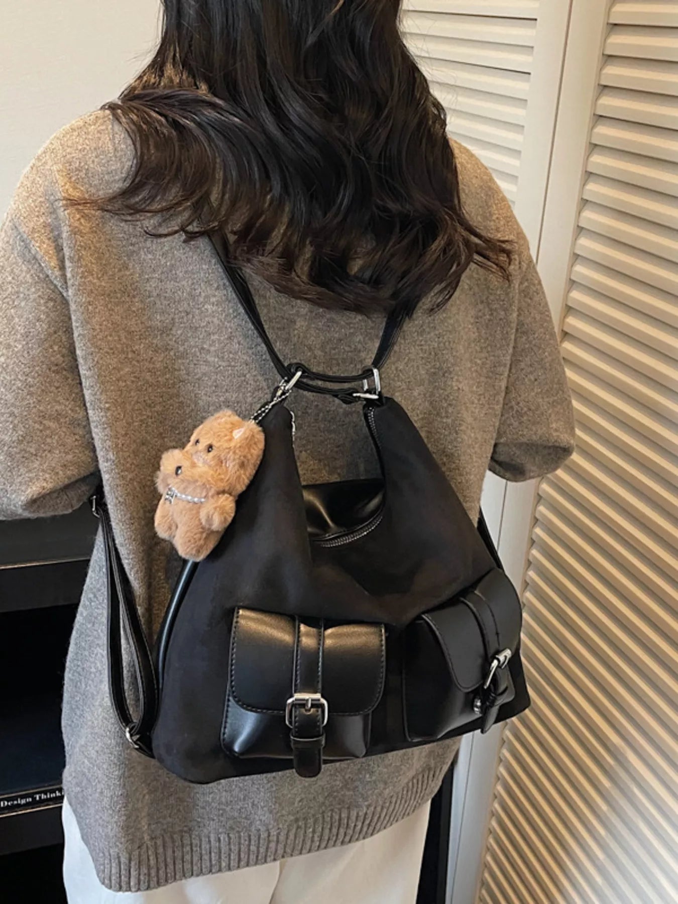 Person wearing a black backpack with a teddy bear charm, standing against a neutral background.