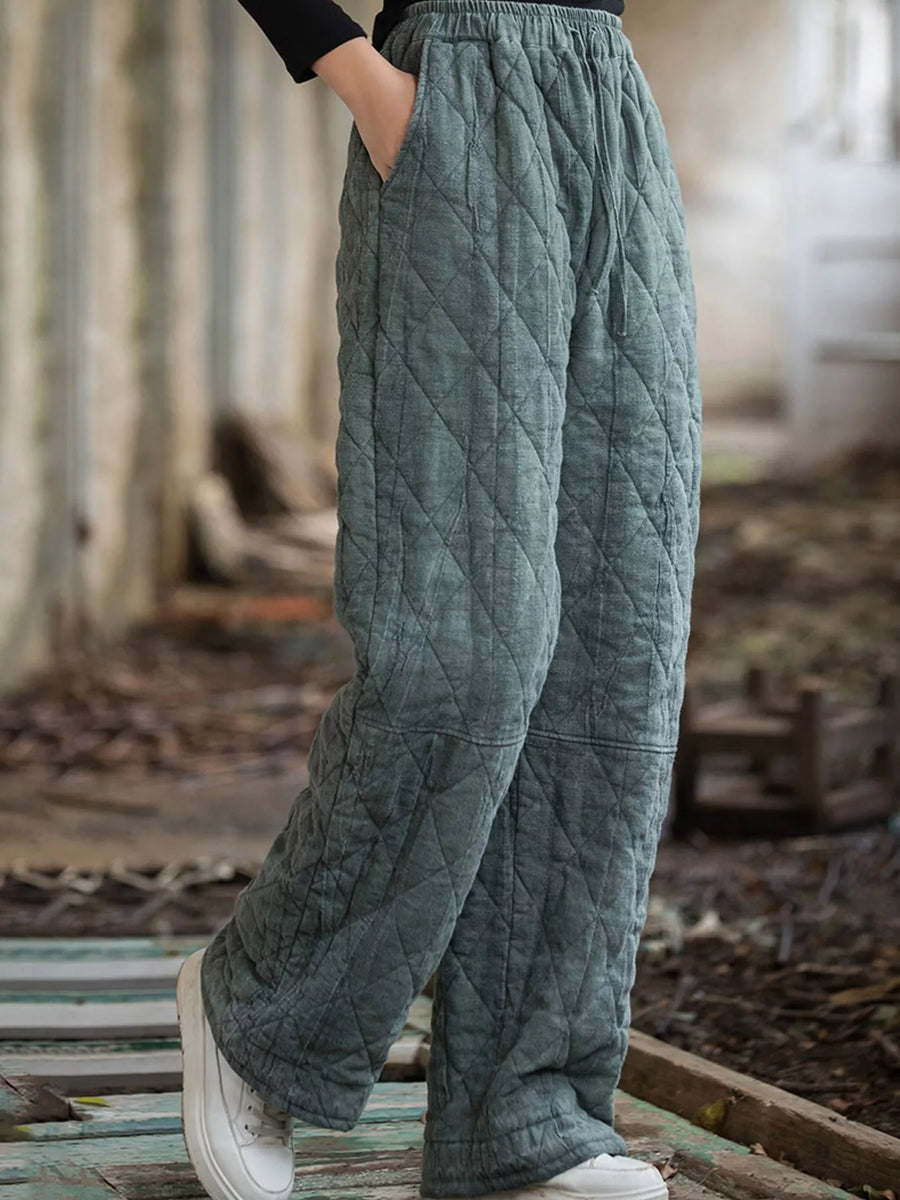A person wearing quilted wide-leg pants in a muted sage green color, standing outdoors on a weathered wooden surface with a rustic, blurred background.