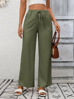 Person wearing green wide-leg pants with a white top and brown sandals, holding a brown handbag against a light gray wall.