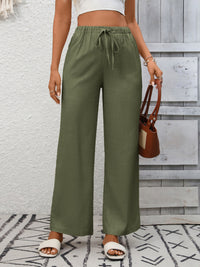 Person wearing green wide-leg pants with a white top and brown sandals, holding a brown handbag against a light gray wall.