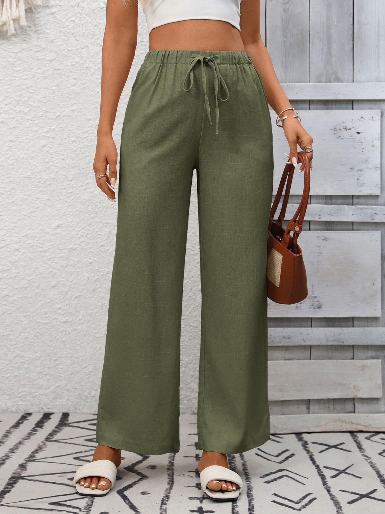 Person wearing green wide-leg pants with a white top and brown sandals, holding a brown handbag against a light gray wall.