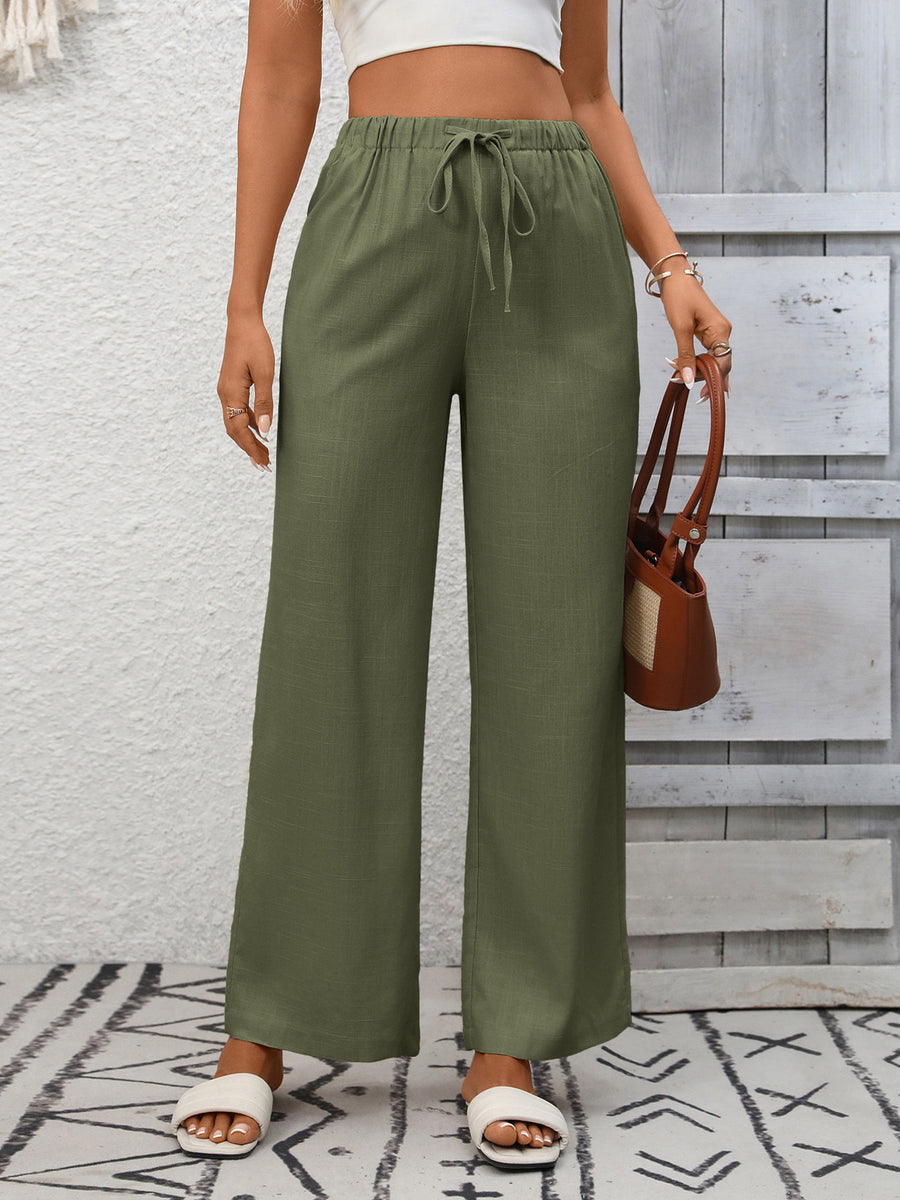 Person wearing green wide-leg pants with a white top and brown sandals, holding a brown handbag against a light gray wall.