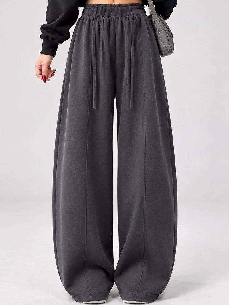 A person wearing dark charcoal gray wide-leg sweatpants with an elastic drawstring waist, styled with a black long-sleeve top and a gray shoulder bag against a light gray studio background.
