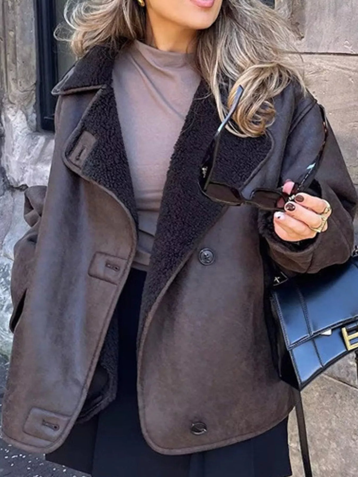 A woman wearing a brown suede-like jacket with a dark brown sherpa lining, styled over a taupe top and black skirt, holding sunglasses and a black handbag.