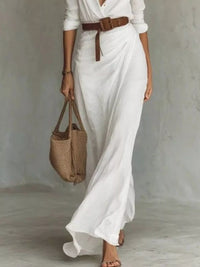 Person wearing a long white dress with a brown belt, holding a woven bag against a neutral background
