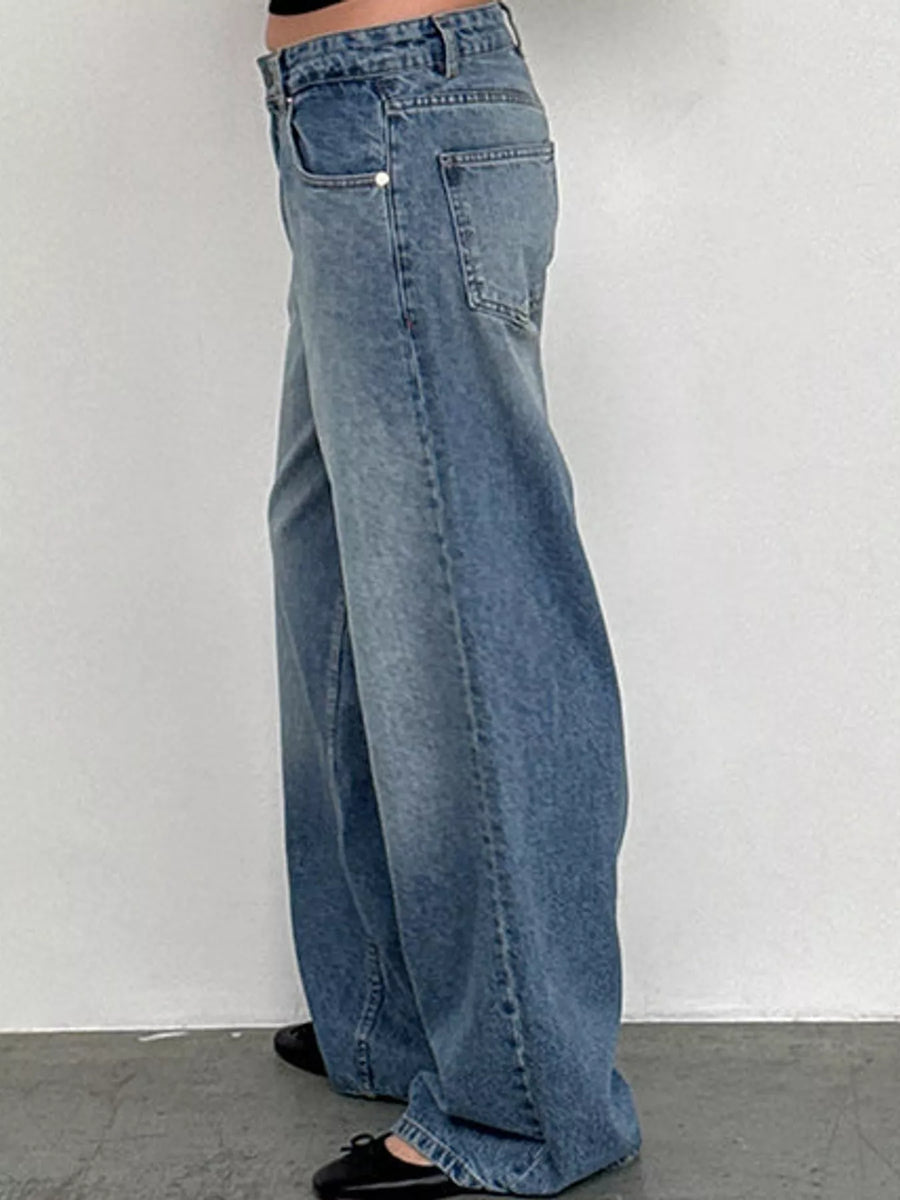 A person wearing wide-leg light blue denim jeans, shown from the waist down against a plain light gray wall and concrete floor.